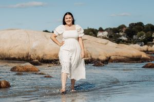 Person in white dress standing among coastal rocks and shallow ocean water.