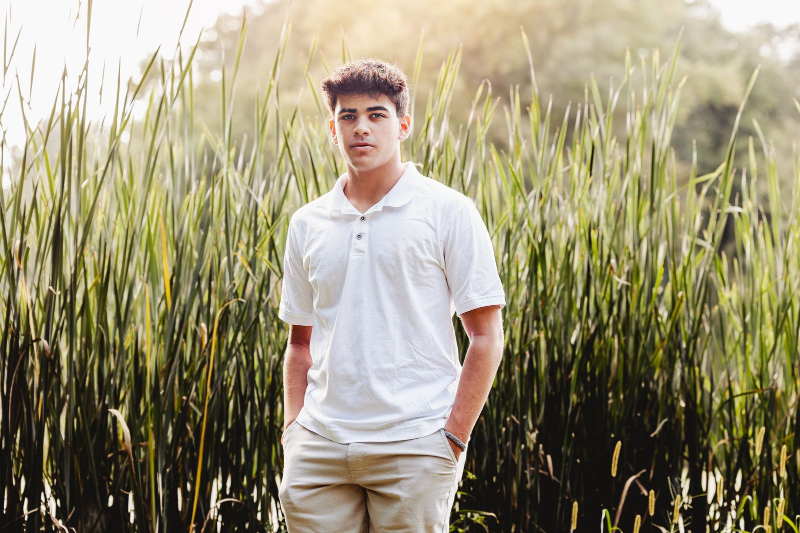Person in white polo shirt poses in front of tall grass during golden hour outdoor photoshoot.