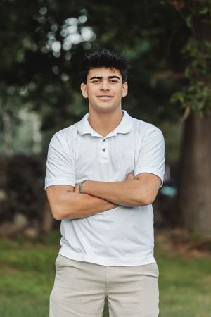 Person in white polo and khaki pants stands with crossed arms against blurred natural background.