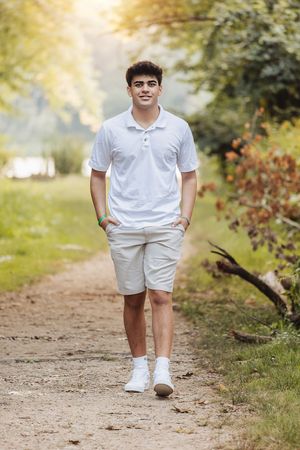 Person in white polo and khaki shorts standing on nature trail with sunlit trees in background.