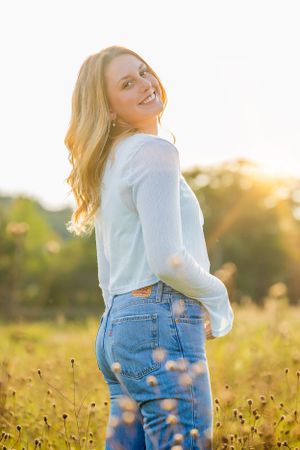 Woman in casual attire poses in sunlit meadow with wildflowers.
