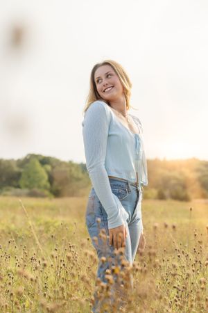 Woman in light blue top and jeans standing in golden wildflower field at sunset.