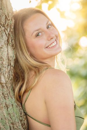 Woman leaning against tree trunk with warm sunset glow in background.
