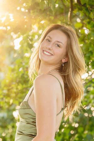 Woman in olive green dress posing against sunlit foliage background.
