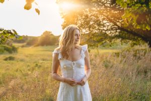 Woman in flowing white summer dress stands in tall grass during golden hour.