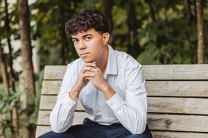 Person sitting thoughtfully on wooden bench in casual attire against natural outdoor background.