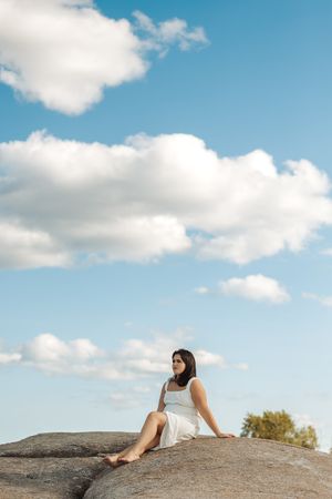 Seated figure in white dress on rock formation against blue sky with fluffy clouds.