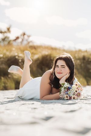 A person in a white dress lying on a beach with a floral bouquet and dreamy sunlit backdrop.