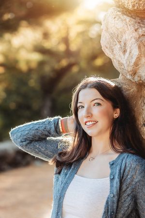 Portrait of someone in a gray cardigan leaning against a tree in warm sunlight with a joyful expression.