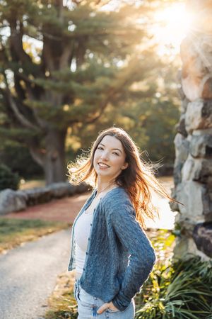 A person stands on a path at sunset, wearing a blue cardigan and jeans while backlit by golden sunlight through trees.
