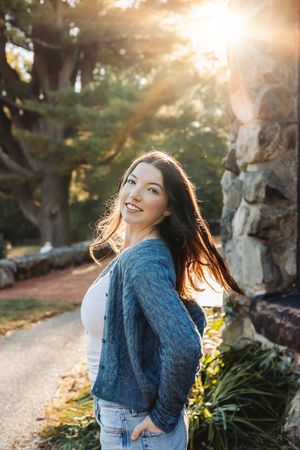 A person in denim and a blue scarf laughs joyfully in golden sunlight while standing outdoors near trees.