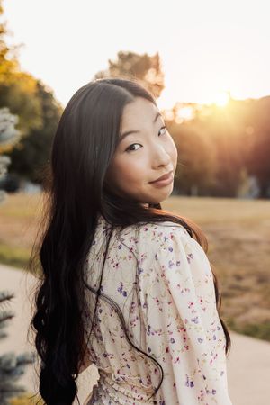 A person in a floral dress poses playfully at sunset in a golden-lit outdoor setting.