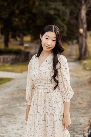 A person in a white floral dress poses outdoors in a park setting during autumn.