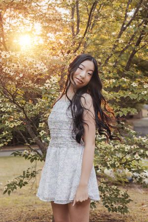 A person in a white lace dress poses joyfully under autumn trees during golden hour sunlight.