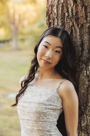 A person in a white floral dress poses against a tree trunk in a natural outdoor setting with soft lighting.