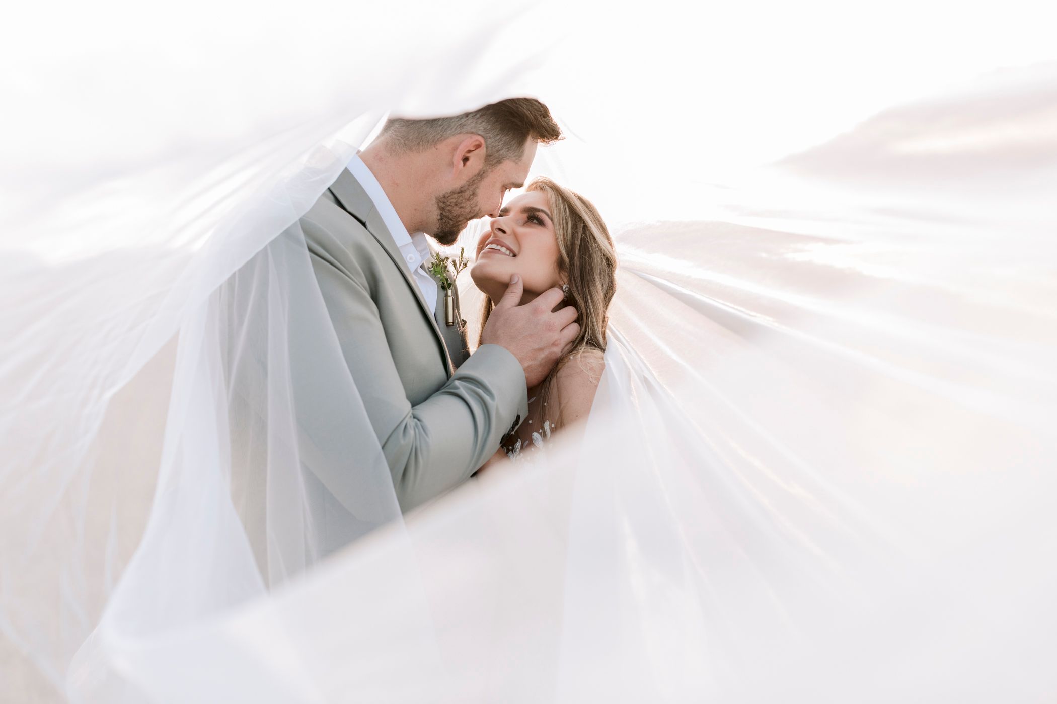 Bride and groom sharing a romantic moment under a billowing white veil in a bright, airy outdoor setting.