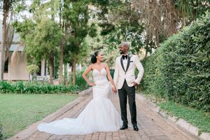 Bride in fitted mermaid gown and groom in white tuxedo jacket holding hands on a brick garden path.
