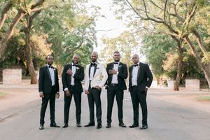 Groom in white tuxedo posing with four groomsmen in black tuxedos on a tree-lined outdoor avenue.