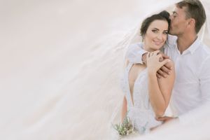 Romantic couple embracing under a flowing white veil, bride holding a small bouquet in a dreamy soft-light setting.