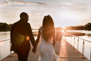 Newlywed couple holding hands walking on a dock at golden sunset during their waterfront wedding celebration.
