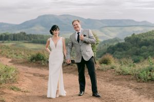 A bride and groom hold hands on a dirt road with scenic mountain views in the background.