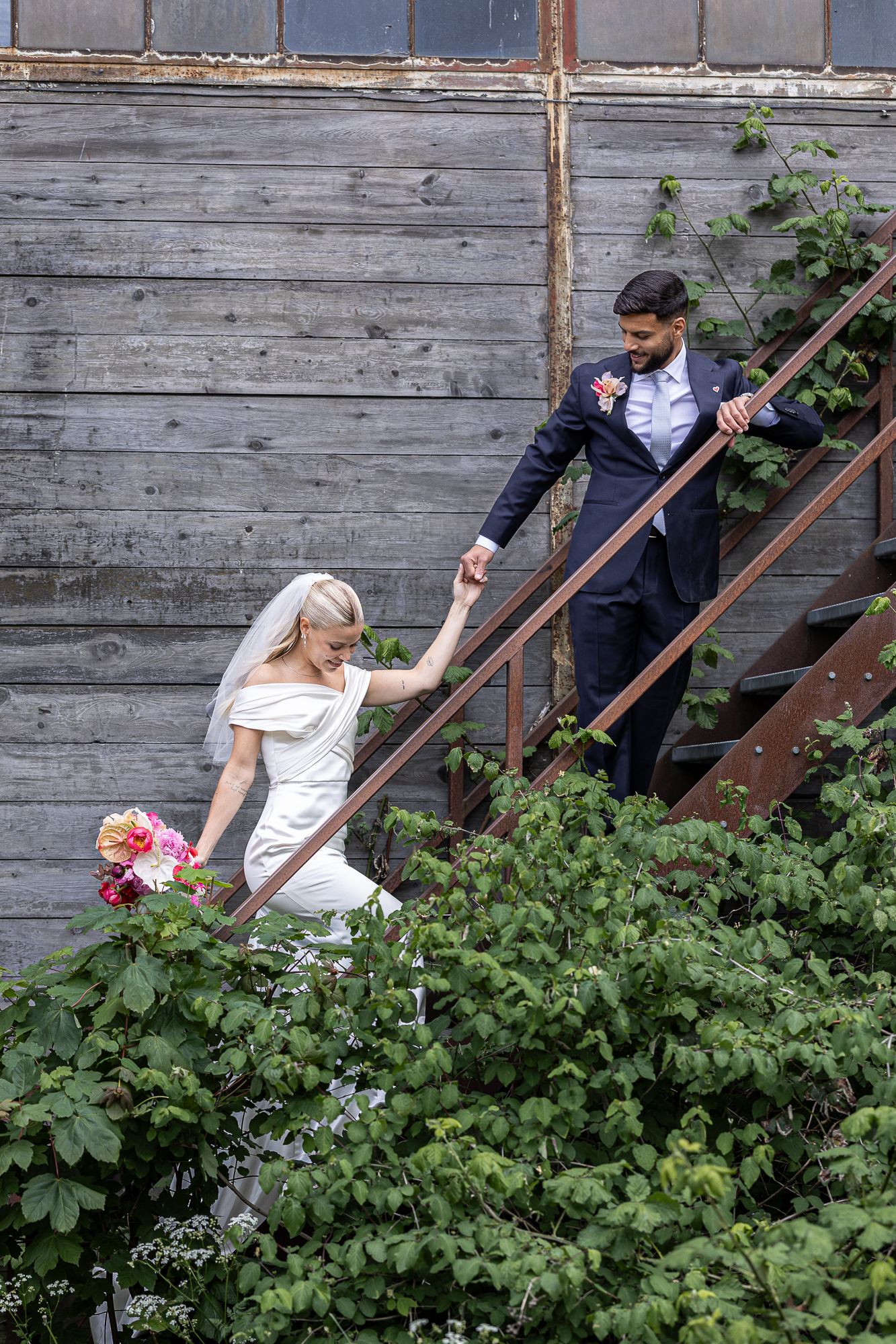 Couple in wedding attire descending rustic wooden stairs surrounded by lush greenery at a rural venue.