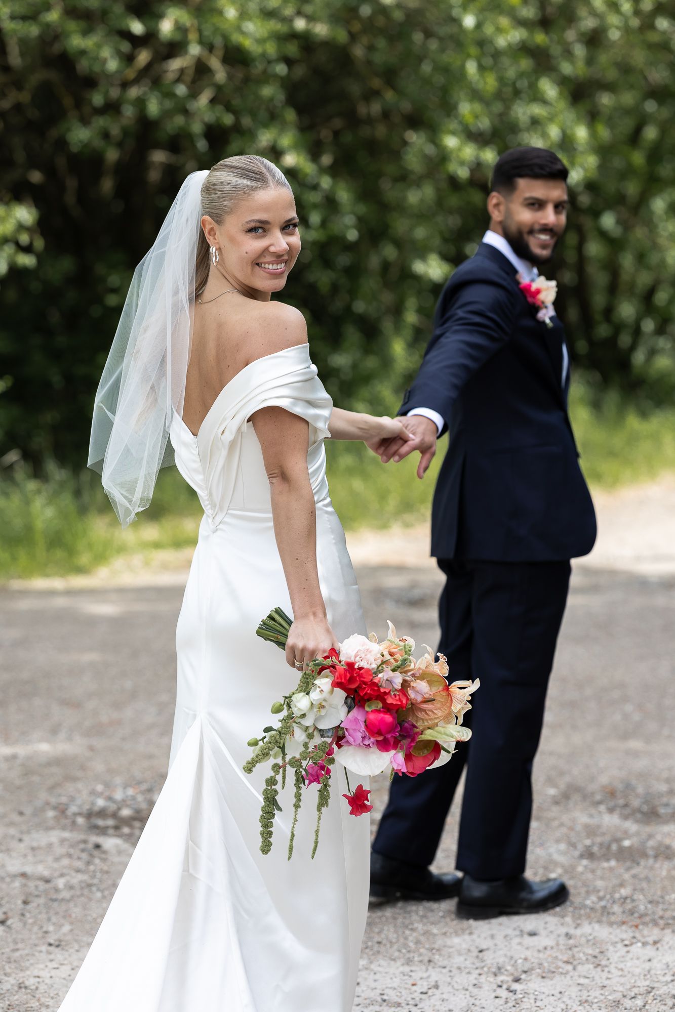 A bride in a white off-shoulder dress holds a colorful bouquet while looking over her shoulder on a wooded path.