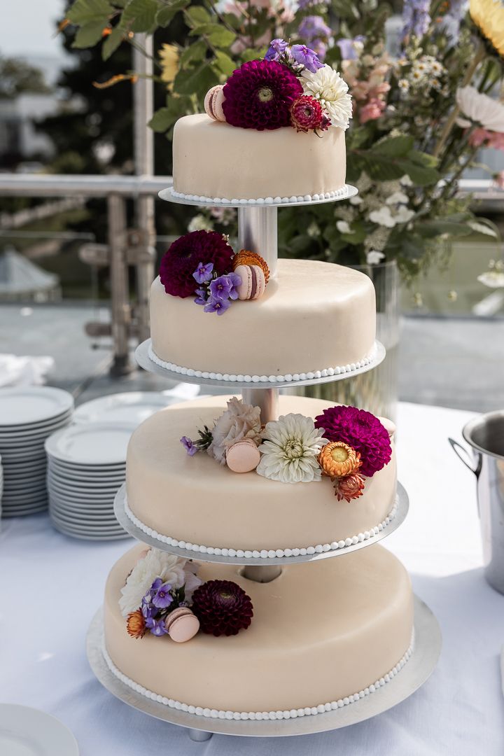 Four-tier white wedding cake decorated with deep purple and pastel flowers on elegant stand.