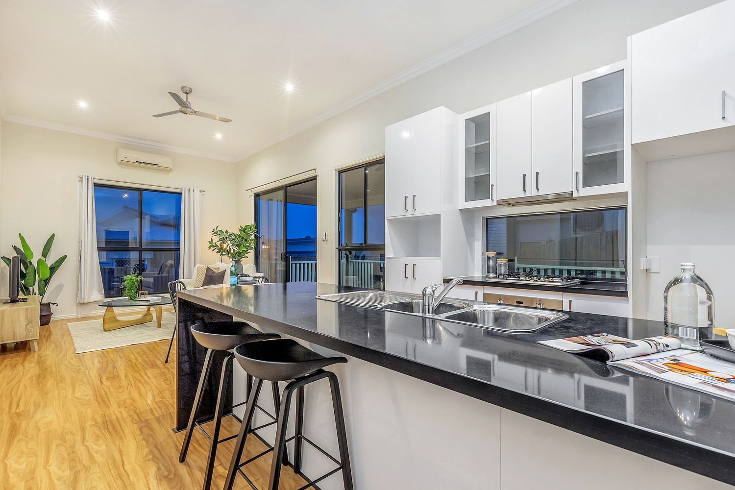 Sleek white kitchen featuring black granite benchtops, bar stools, and open-plan living area with city views.
