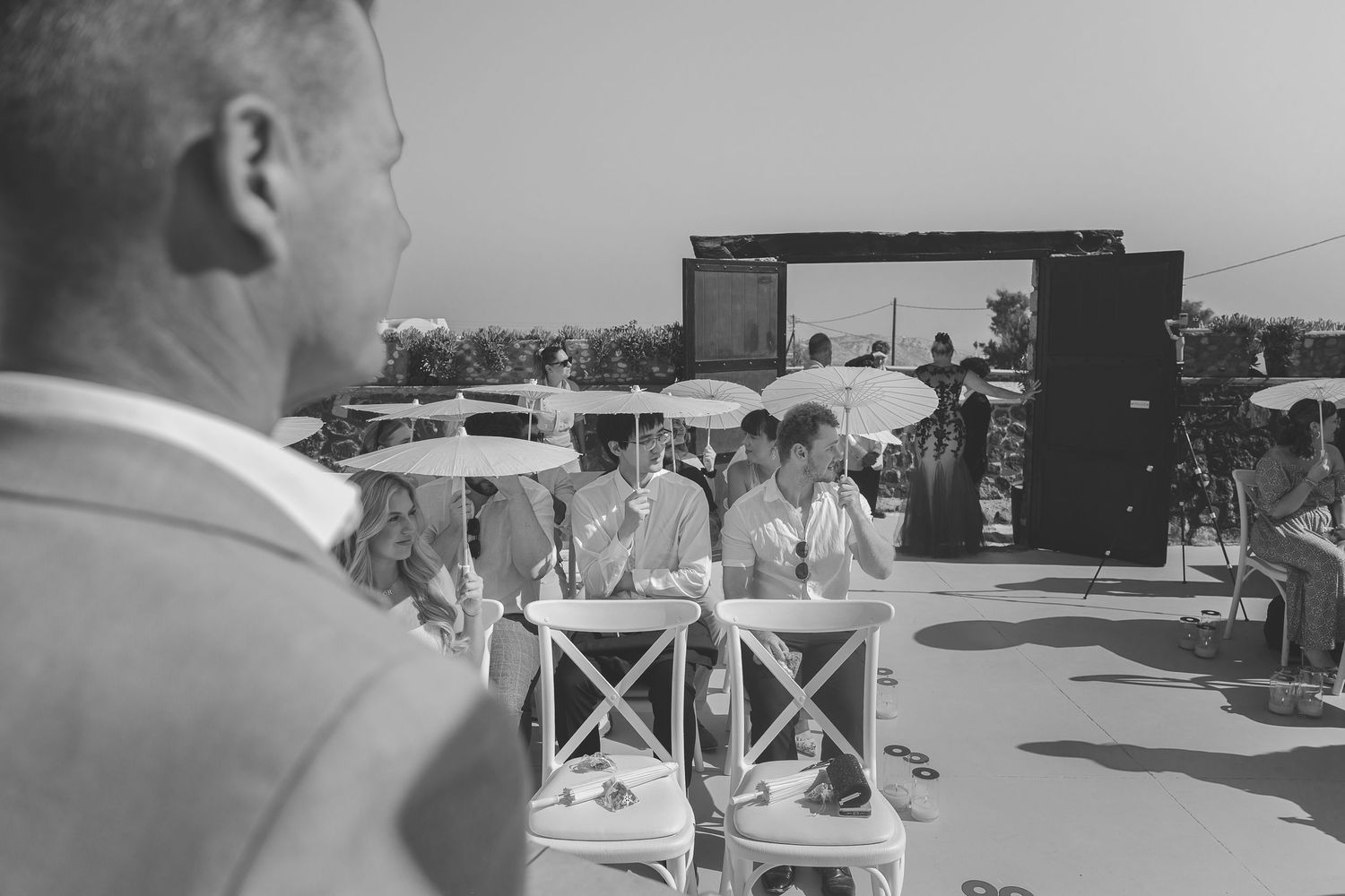 A view over the shoulder of a destination wedding celebrant of the guests assembled on chairs with parasols at El Viento in Santorini.