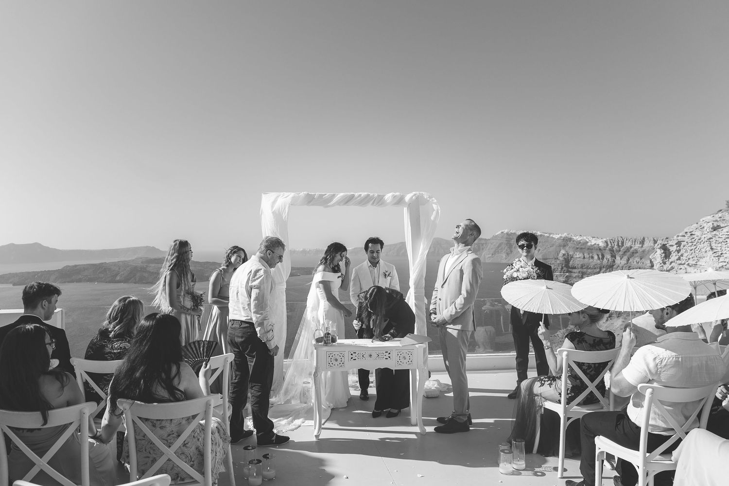 A black and white photo of an outdoor Santorini wedding ceremony with chairs and guests gathered on a sunny day at El Viento