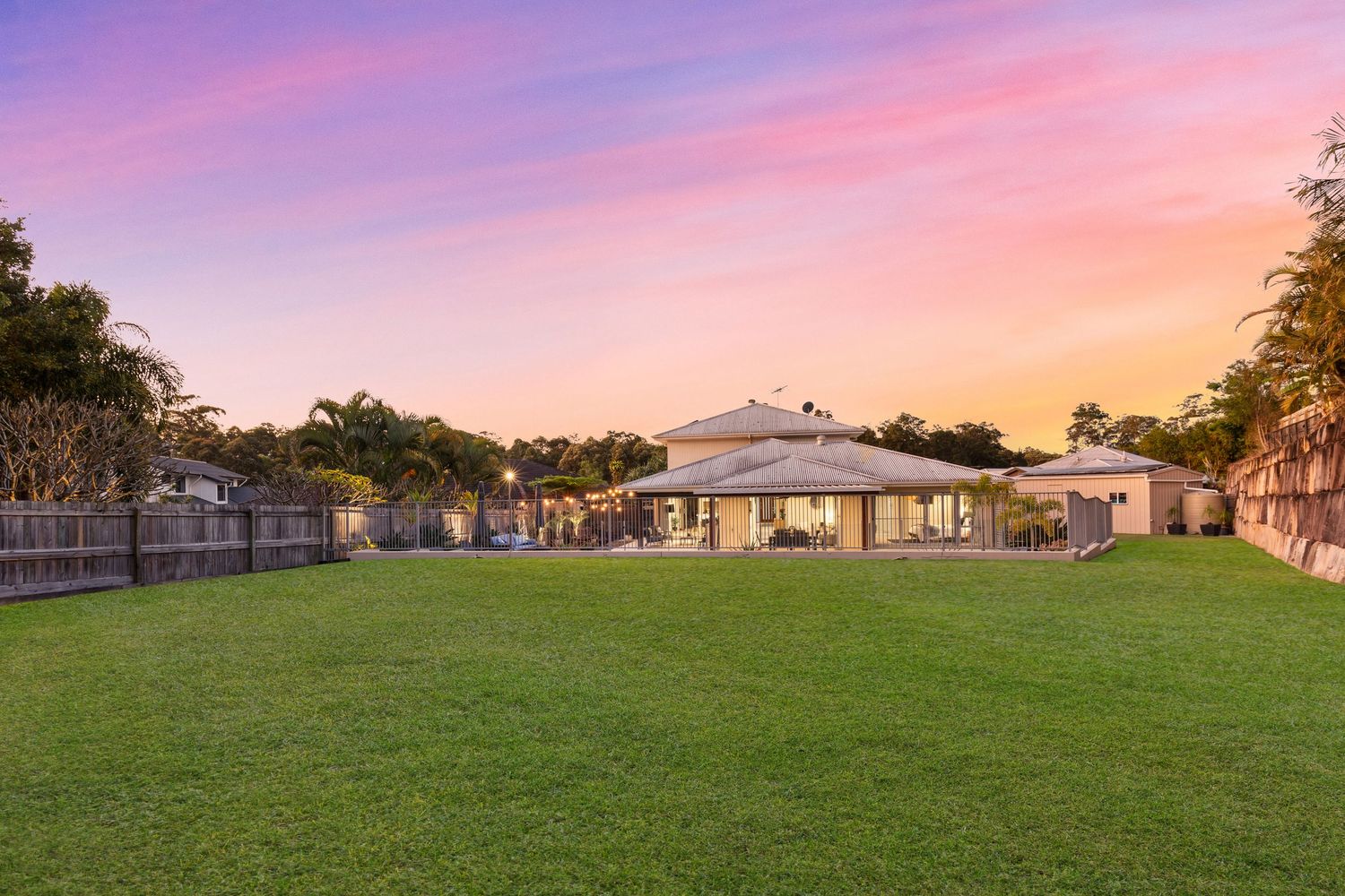 Spacious fenced backyard with manicured lawn, tropical trees, and a cozy home illuminated at dusk.