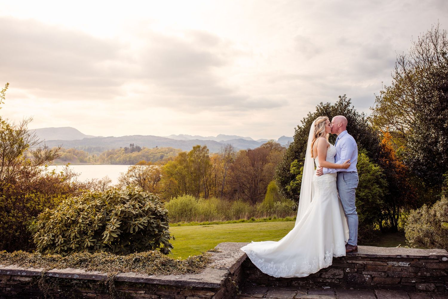 A wedding couple stands in a scenic mountain landscape at sunset with dramatic clouds in the background.