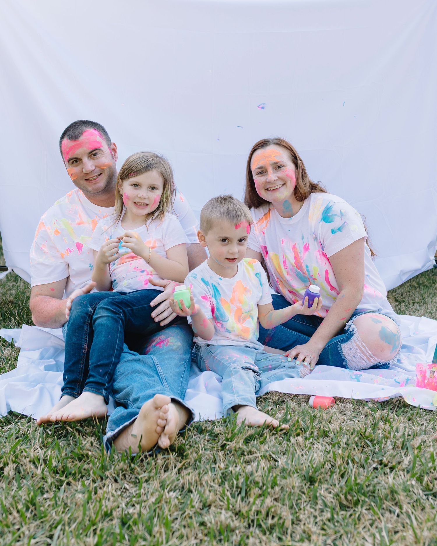 A family wearing white t-shirts with colorful paint splatters sits together on grass during an outdoor photo session.