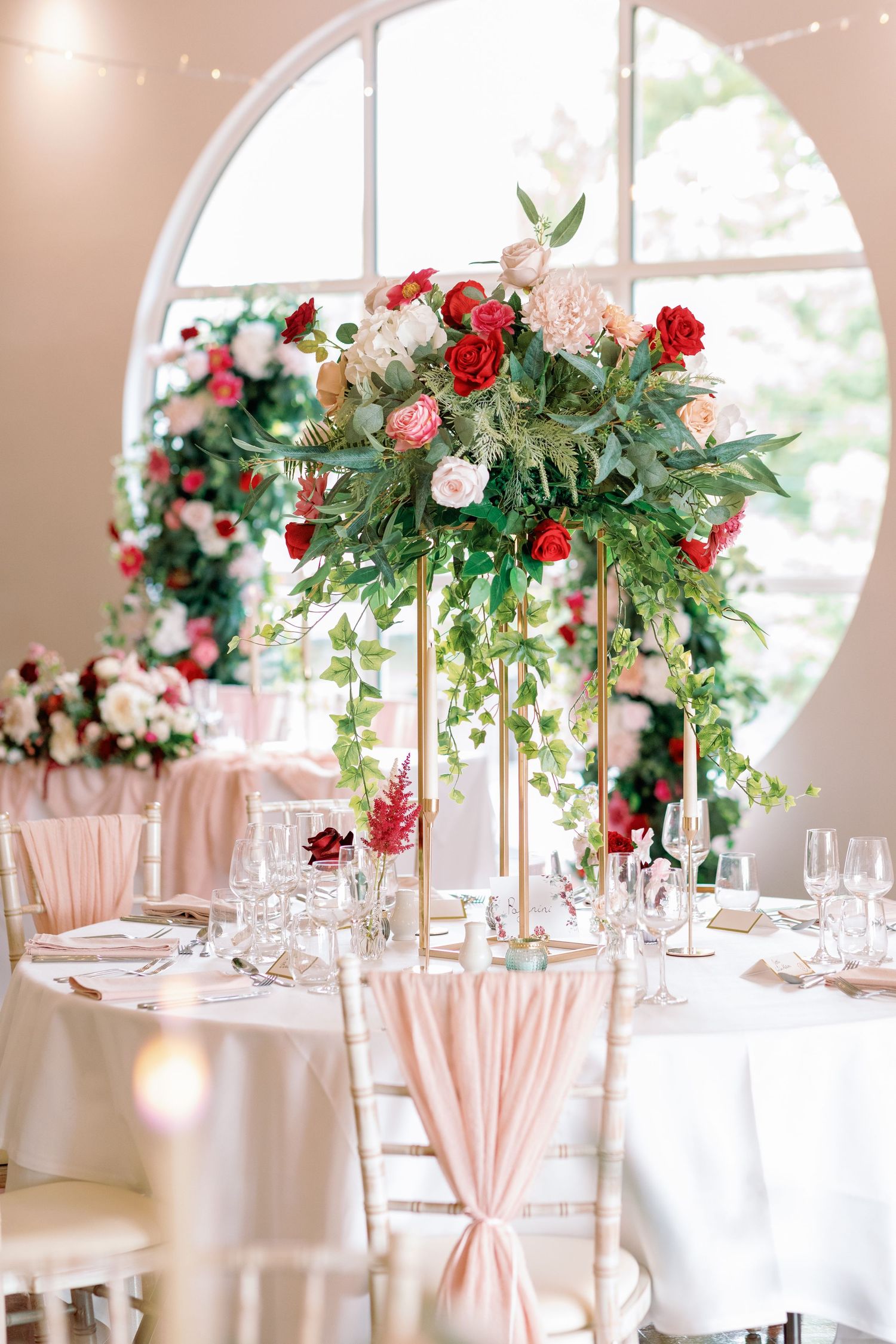 Elegant wedding reception table with tall floral centerpiece featuring red roses and greenery in a bright venue with arched window.
