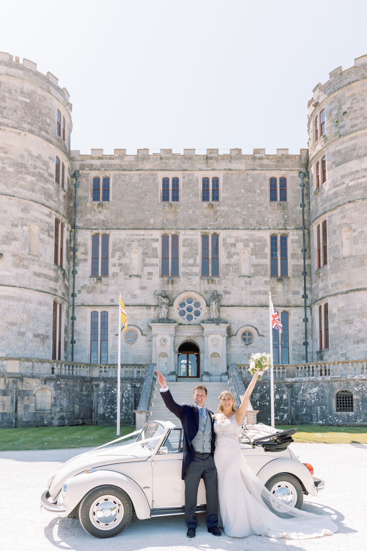Classic white vintage car parked in front of a majestic stone castle with crenellated walls and towers.