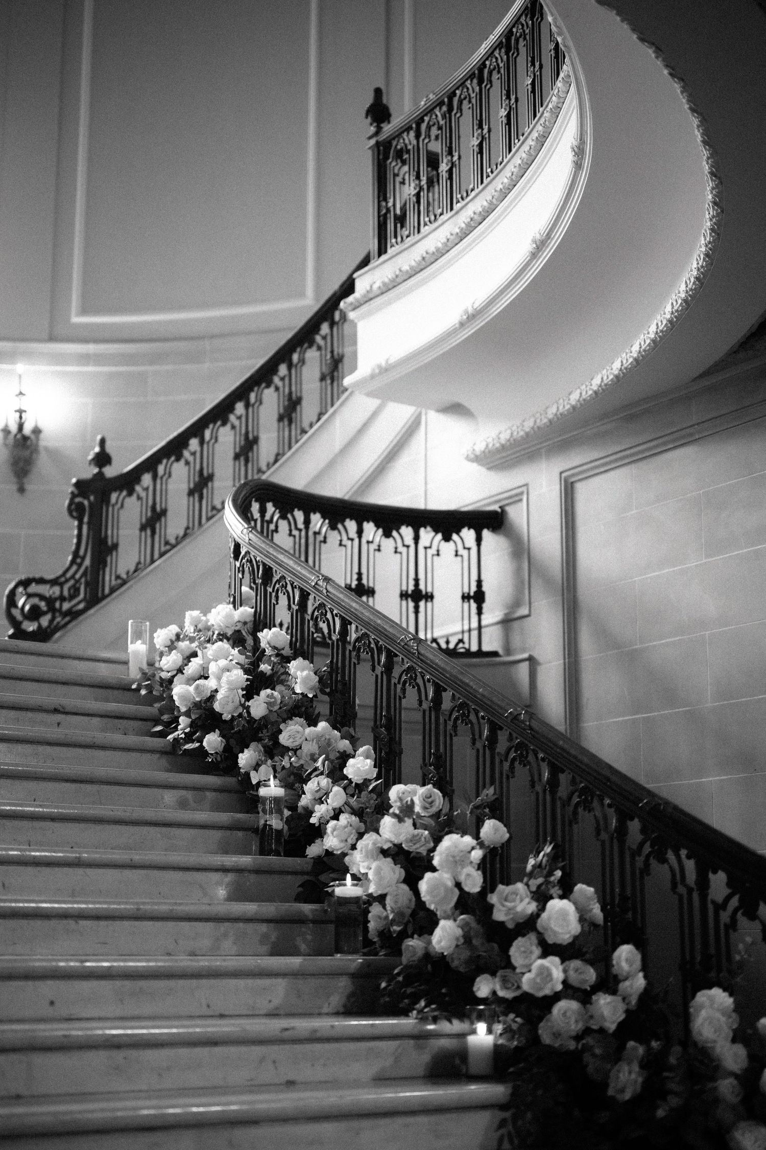 Elegant curved staircase with ornate black iron railings decorated with white roses in black and white photography.