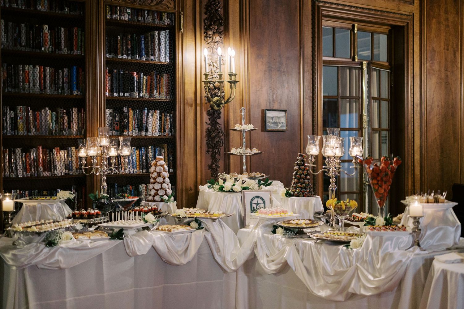 Elegant wedding reception buffet table with white linens set against wood-paneled library walls with candlelit sconces.