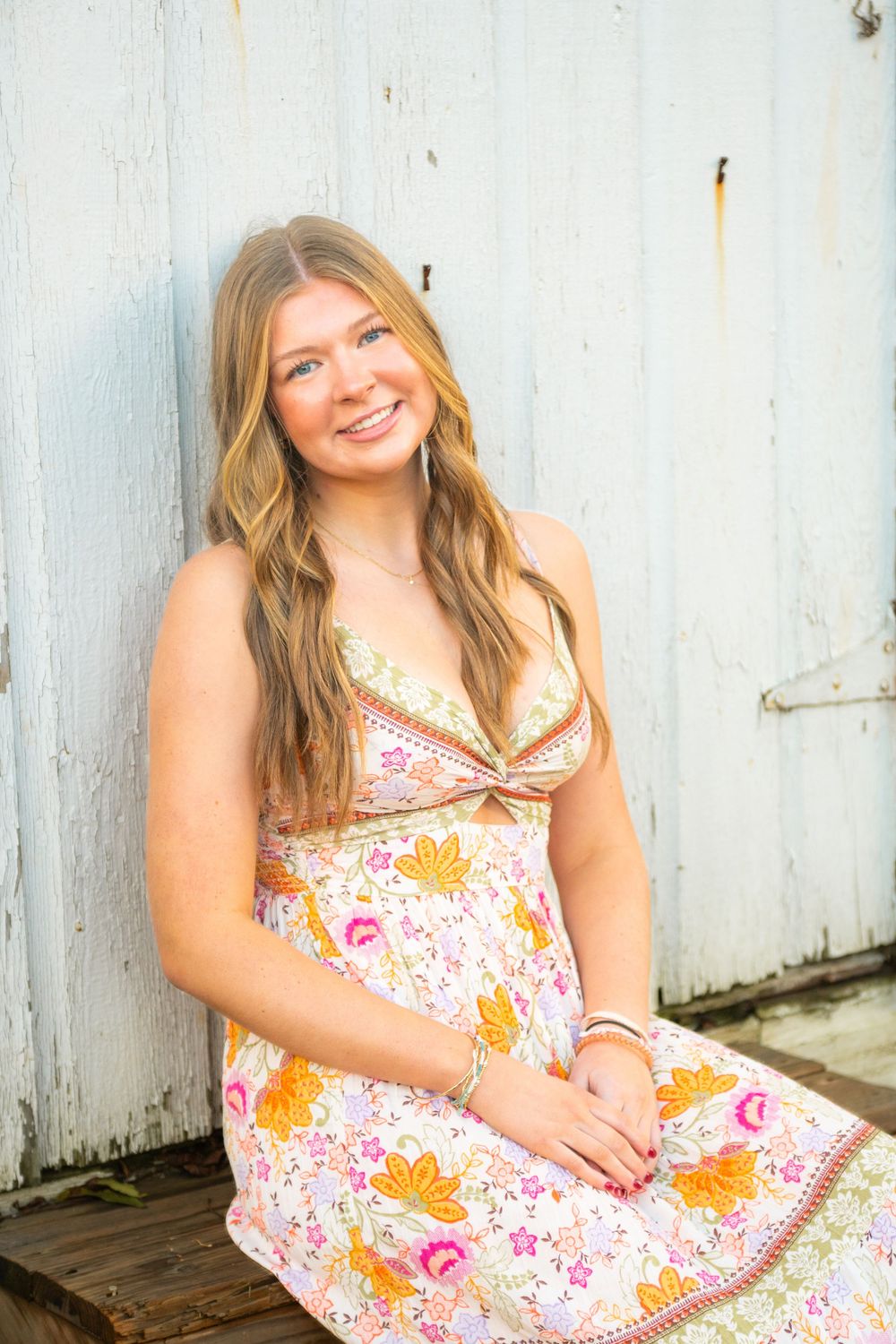 A floral sundress with pink and orange patterns is worn while posing against a white wooden wall background.