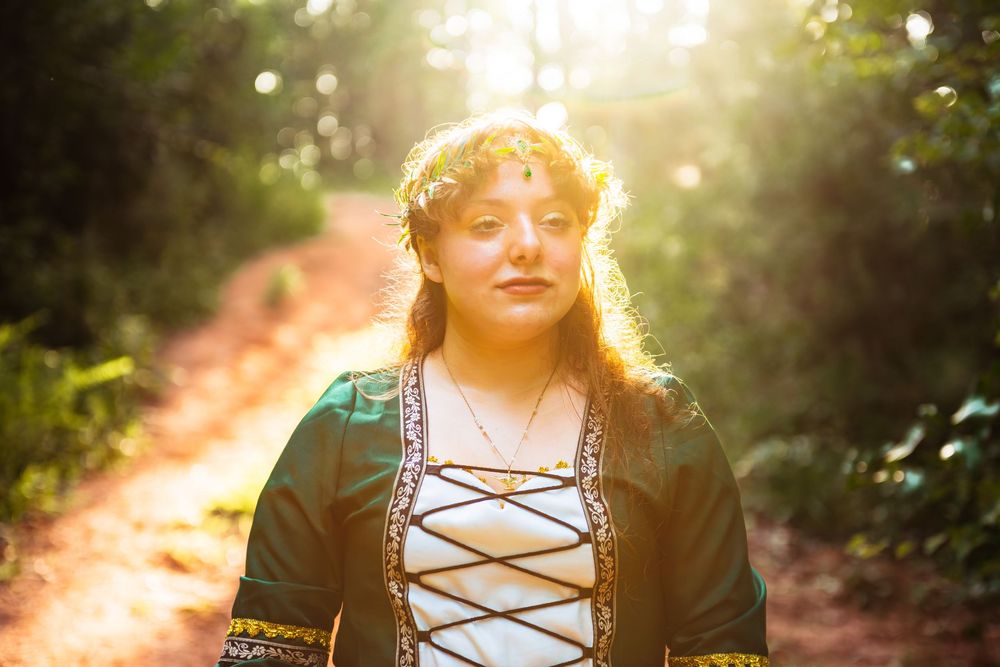 A person in medieval renaissance style dress poses in golden sunlit forest during a fantasy photoshoot.