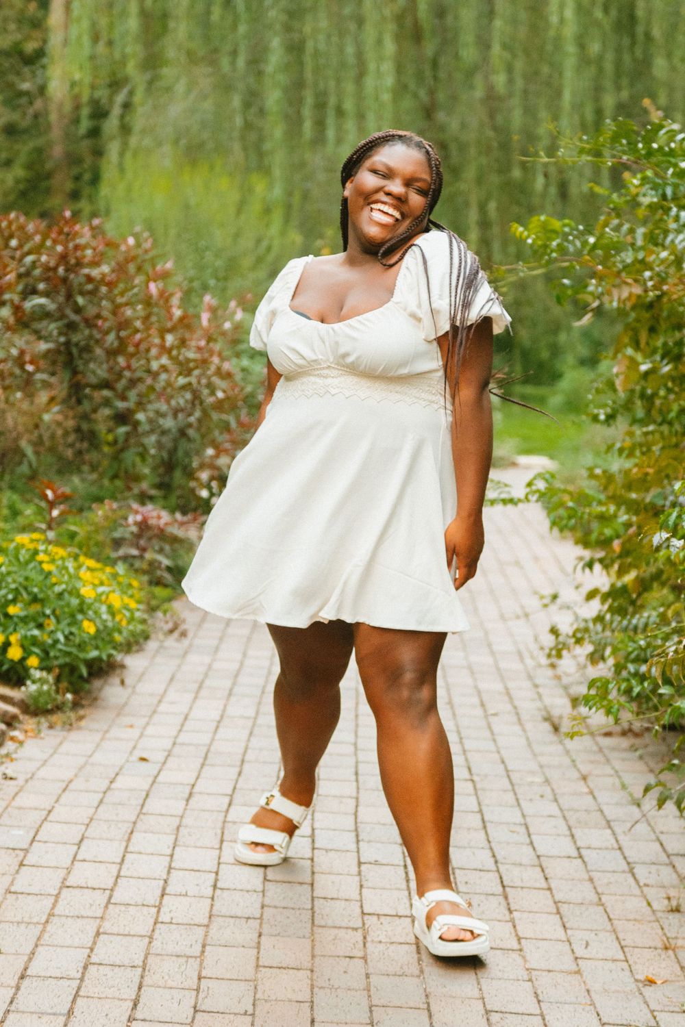 A joyful person wearing a white summer dress and sandals poses on a brick path surrounded by lush garden foliage.