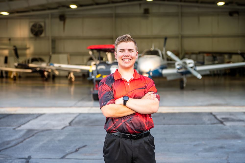 Person in red uniform stands with arms crossed in an aircraft hangar with planes visible in the background.