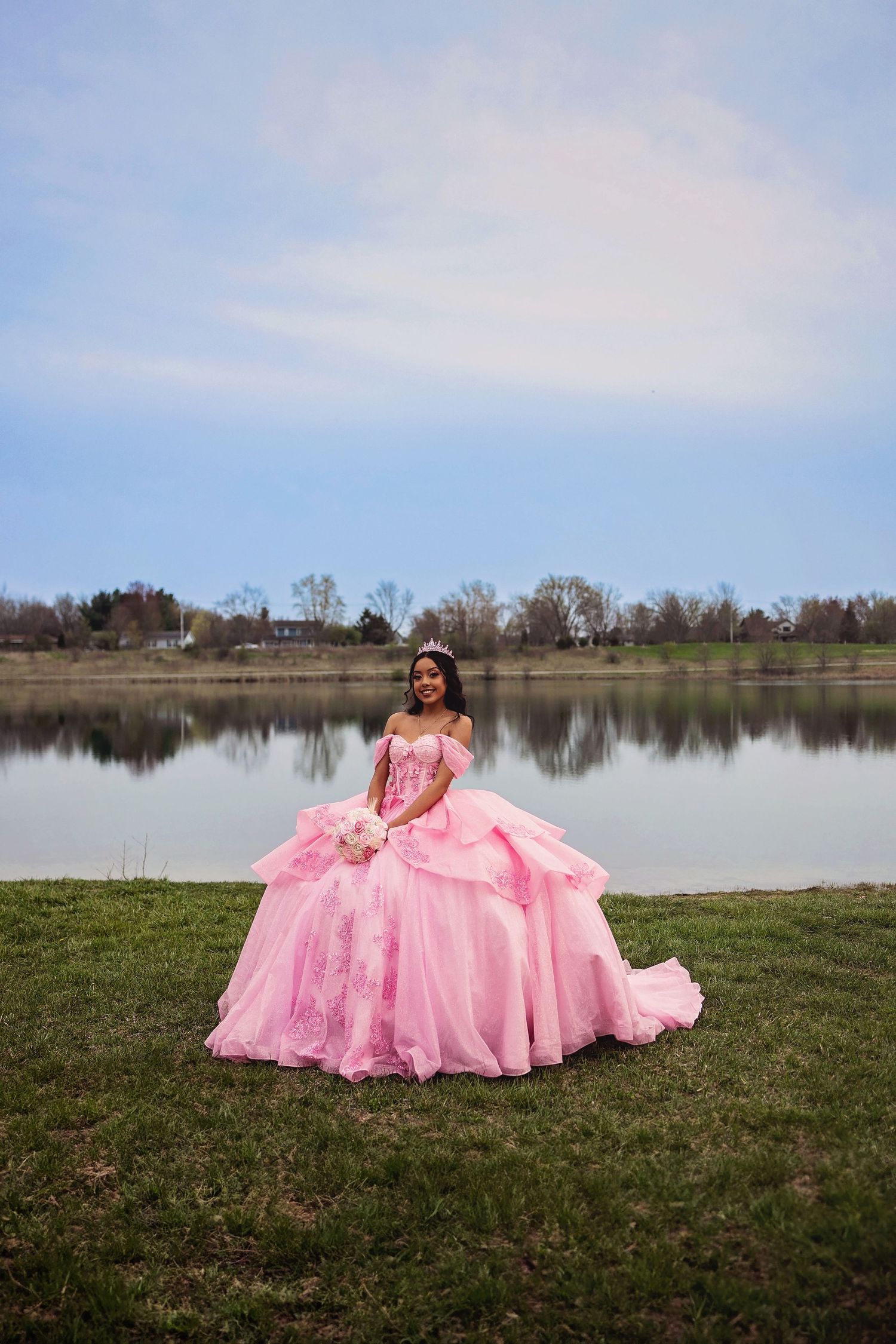 A pink ballgown flows across green grass near a serene lake under blue skies.