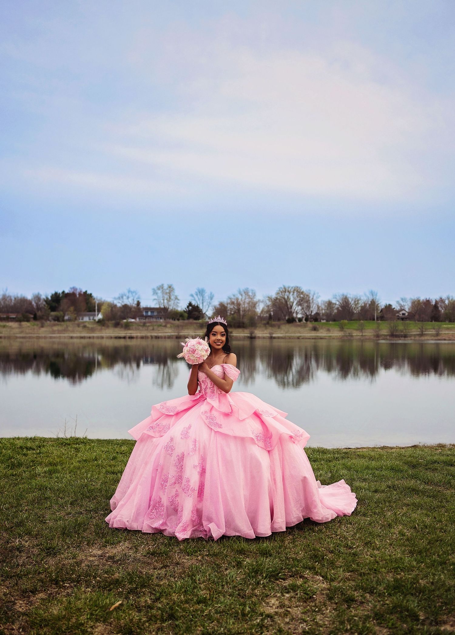 A stunning pink quinceañera dress spreads across grass by a tranquil lake.