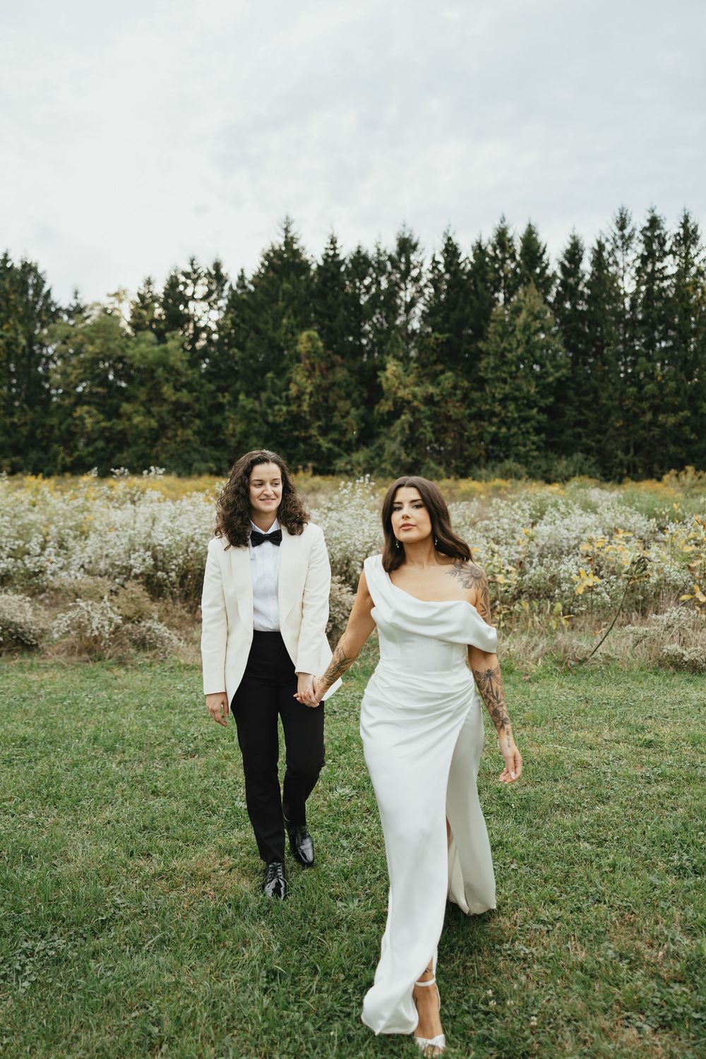 A couple in wedding attire walks together through a wildflower meadow with evergreen trees in the background.