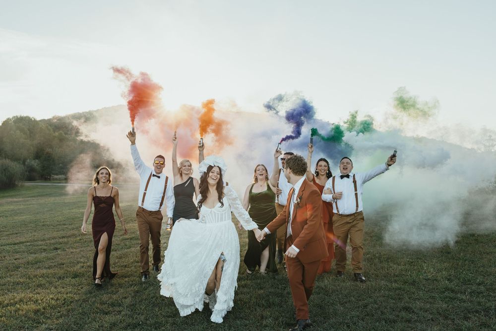 Wedding party runs through a field with colorful smoke bombs creating a vibrant outdoor celebration scene.