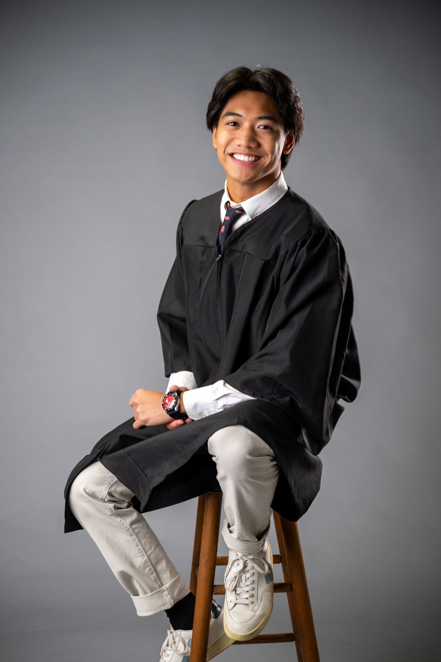 A graduate in a black academic gown and tie  against a gray studio backdrop at Senior portraits at Greenwich High School.