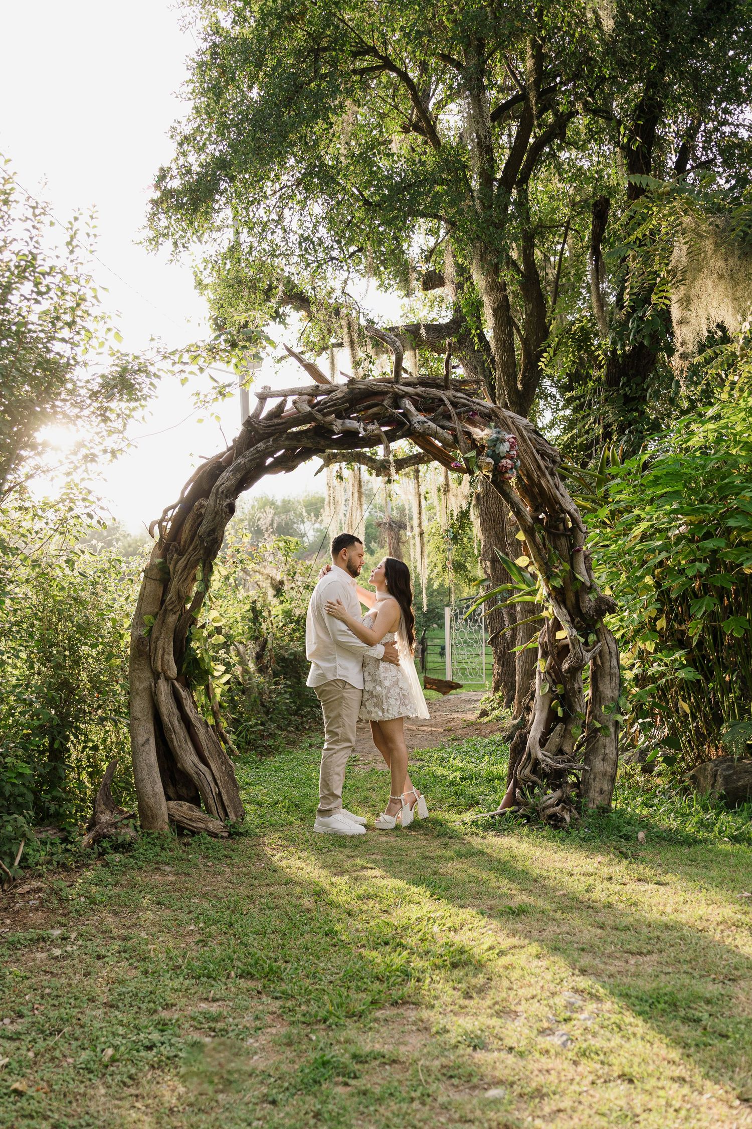 A couple shares a romantic moment among ancient olive trees in a sunlit garden setting during a photo session.