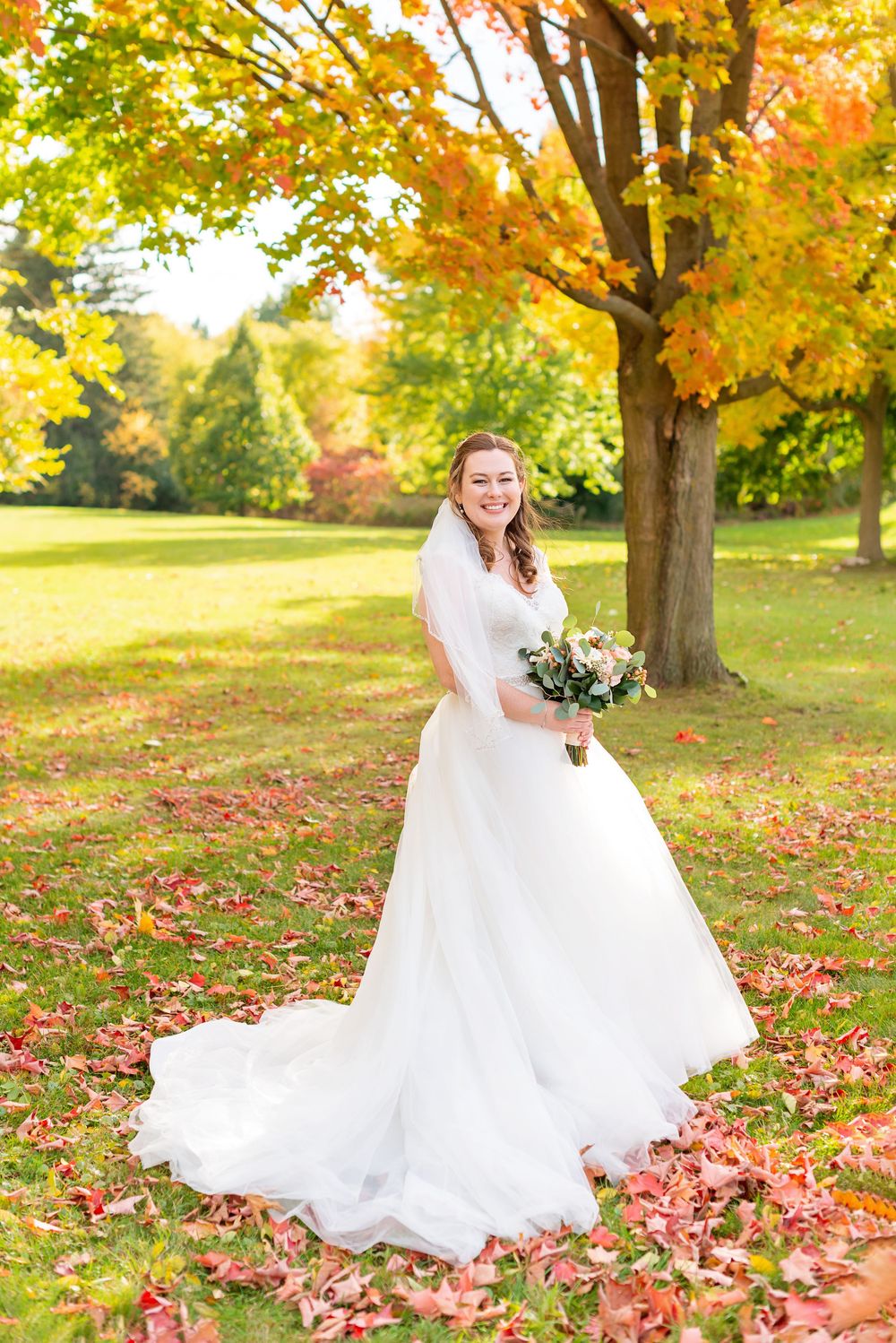 A bride in a flowing white gown stands amid autumn foliage and fallen leaves in a scenic park setting.