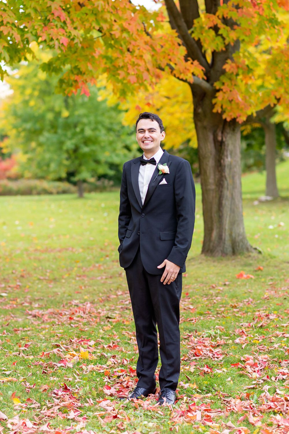 A groom in a black tuxedo poses outdoors among autumn trees with colorful fall foliage and scattered leaves.